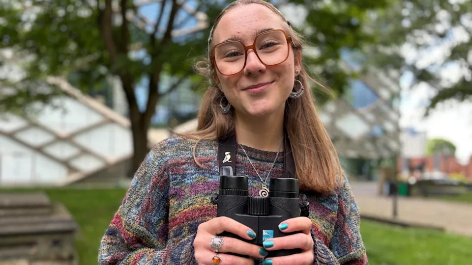A young woman with hair tied back and wearing a colourful jumper, holds a pair of binoculars