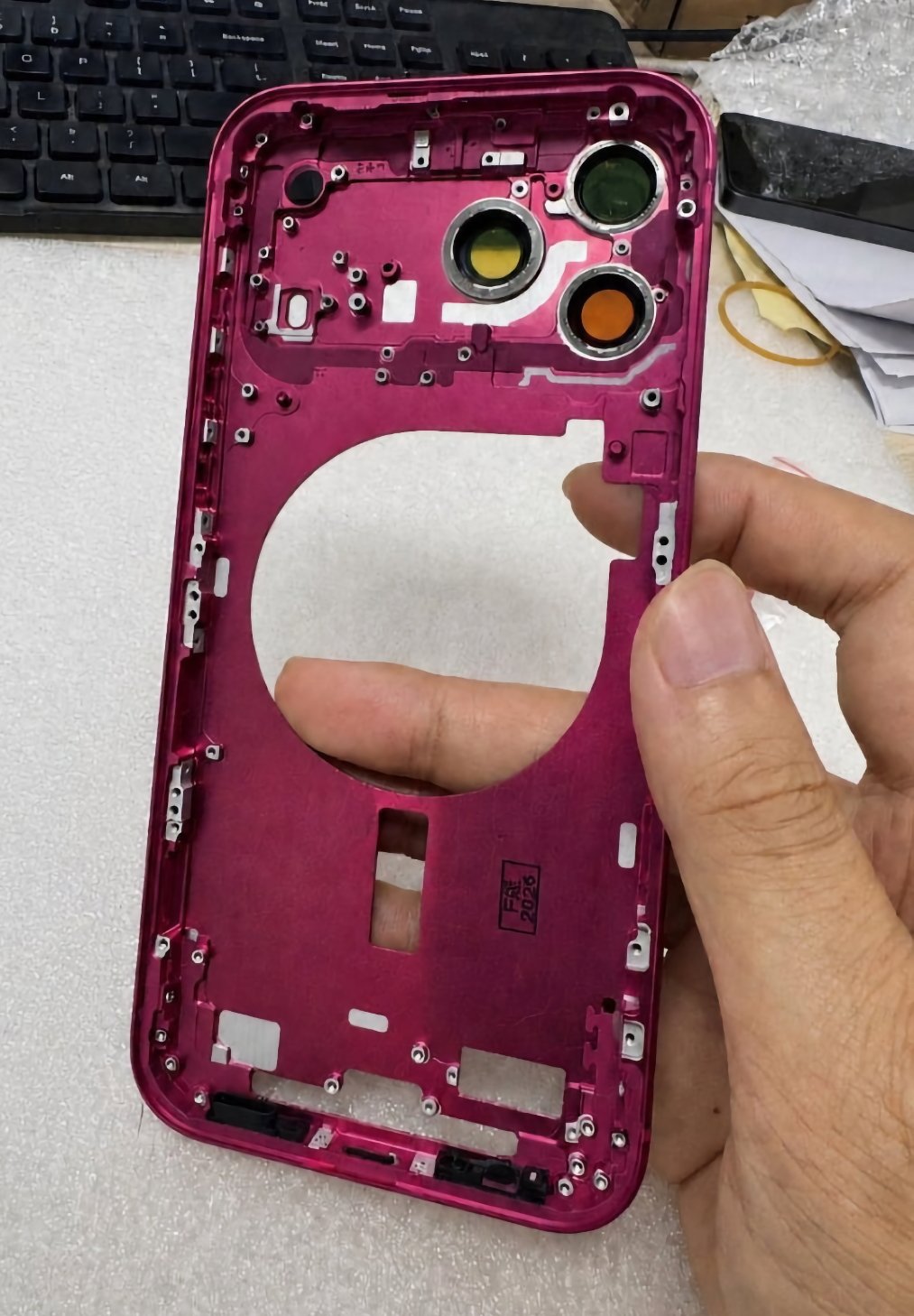 Hand holding a metallic magenta smartphone frame with large circular cutout and three camera lens openings, resting on a light surface near a keyboard and scattered papers