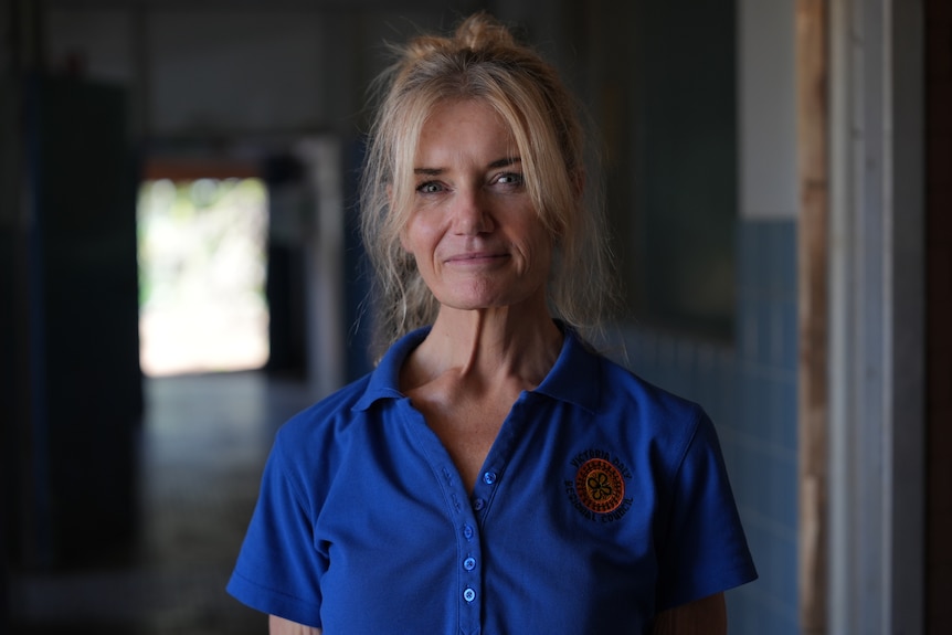 White woman, blonde hair tied up in loose bun, fringe hair, blue polo shirt, standing inside dark lit building.