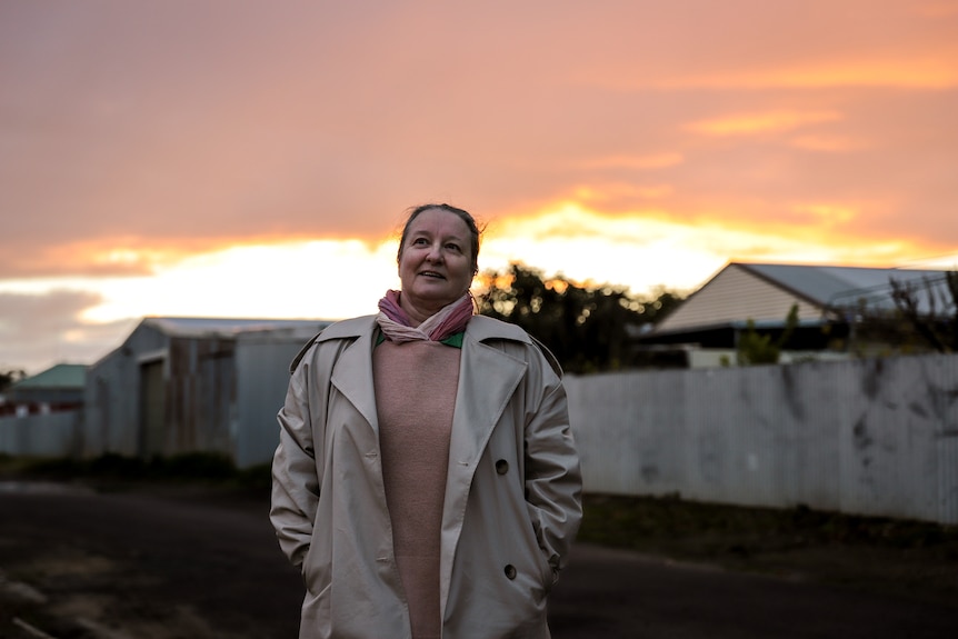 Woman wearing jumper and cream coat stands in country backstreet beneath fading sunset