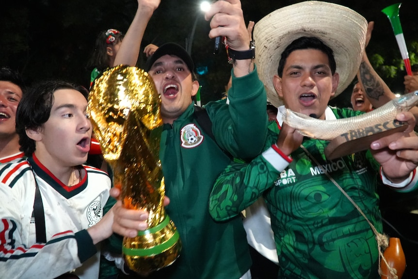 Three male Mexican football fans cheer, one is holding a World Cup trophy.