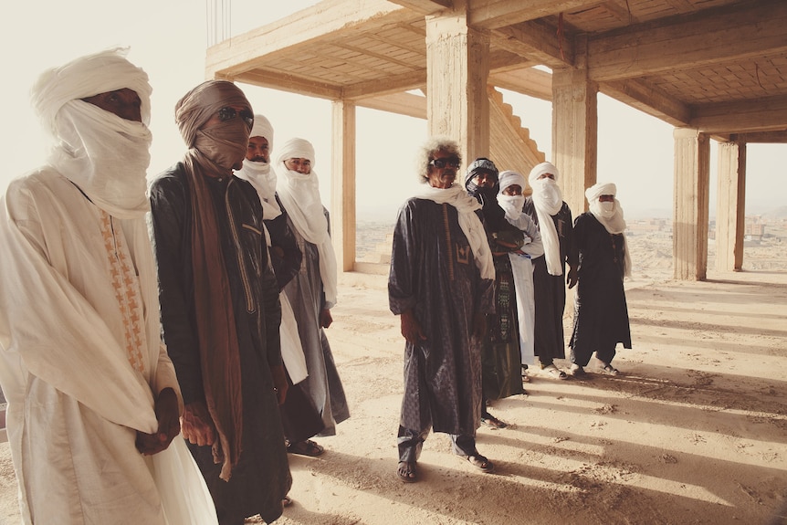 Tinariwen wear headscarves and stand inside a concrete structure overlooking a dusty town.
