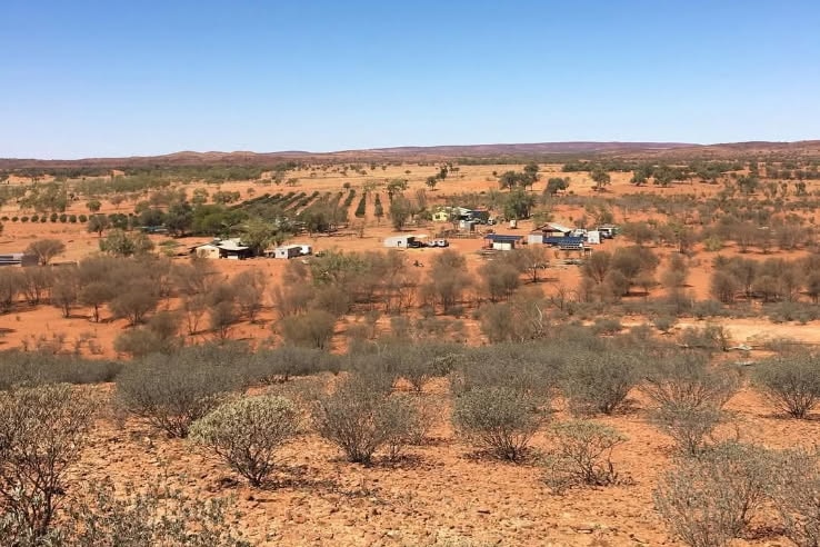 A landscape of orange dirt and low green bushed with a small cluster of houses in the distance