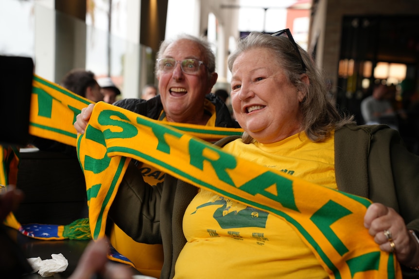 A lader and an older man each hold their Socceroo scraves and smiles, while wearing Socceroo shirts.