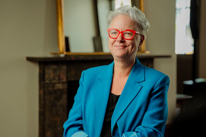 Ms Sharpe sitting in a room on a chair smiling while wearing a blue blazer.