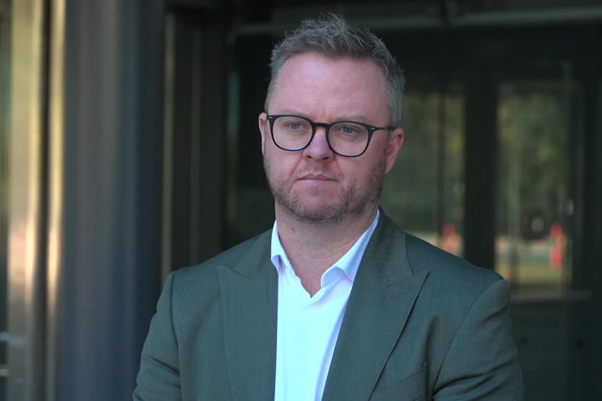 A man in a grey suit and glasses is standing outside a glass building.