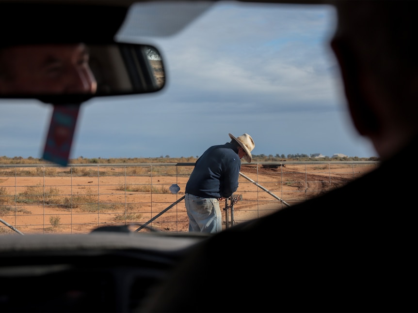 Man wearing weathered hat opens a farming fence on a country property while silohuetted man in a car watches on