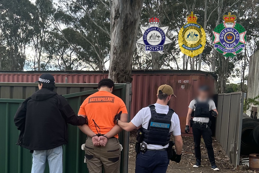 A man in an orange shirt is arrested with his hands behind his back, by three officers. A shipping container is in the backgroun