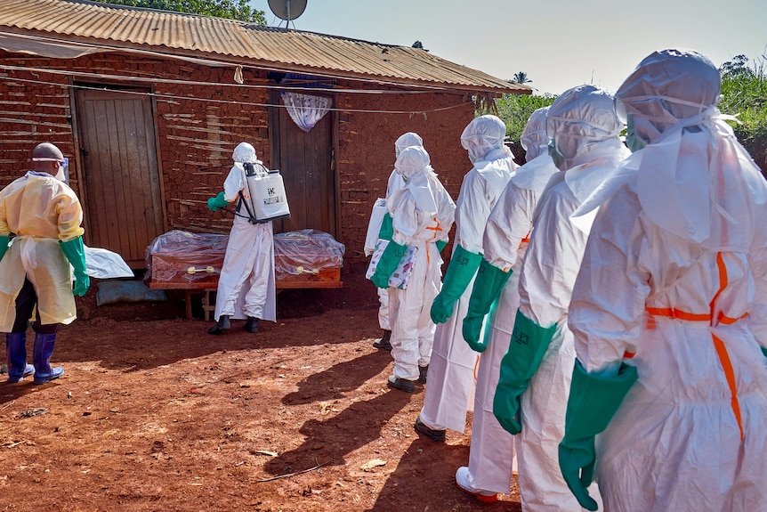 A line of medical workers in full personal protective equipment line up outside a house ready to retrieve a coffin.