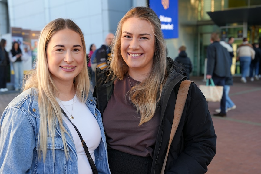 Two people with long blonde hair stand outside smiling, near the stadium.