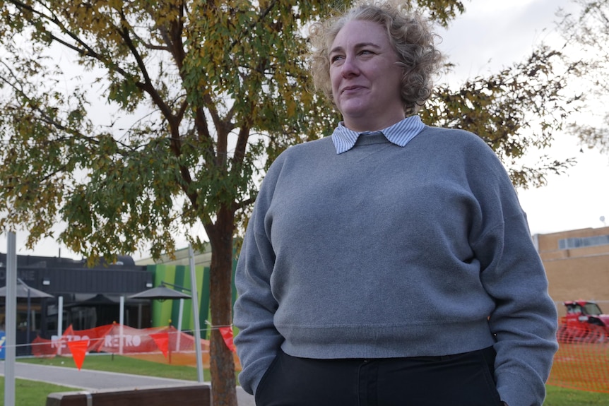 A grey-haired woman stands, hands in pockets, on a public footpath.
