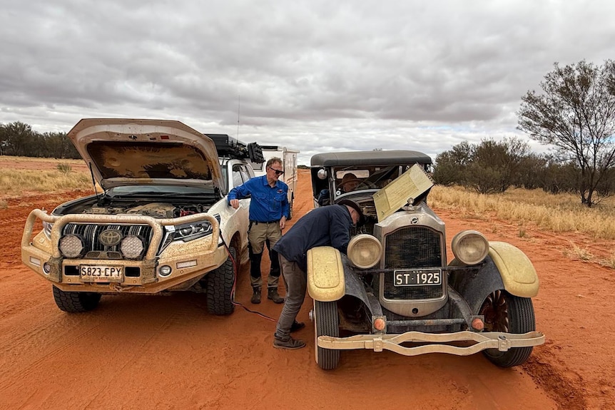A man has his head stuck in the bonnet of a vintage car while cables run to a prado pulled up next to him on the red dirt.