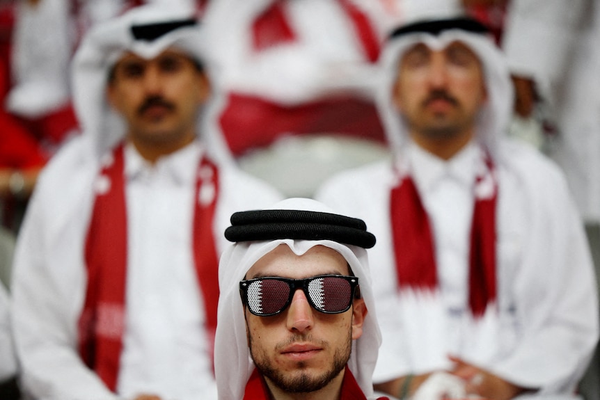 a man wearing a white thobe and headscarfe and sunglasses, while others in the same attire sit behind him