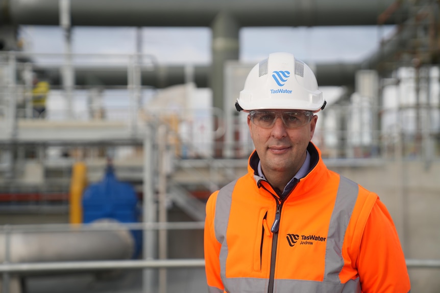 A man wears hi-vis and glasses and stands by sewerage infrastructure