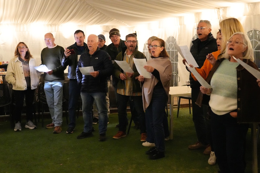 A group of men and women under a white marquee holding piece of paper with lyrics and singing.