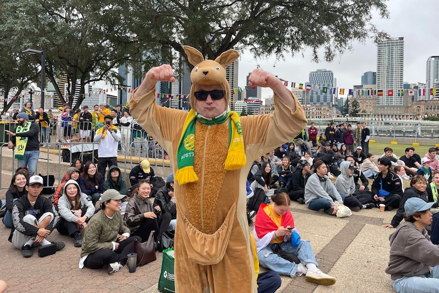 A man wearing black sunglasses and a full-body kangaroo costume with a Socceroos scarf around his neck shows of his muscles.