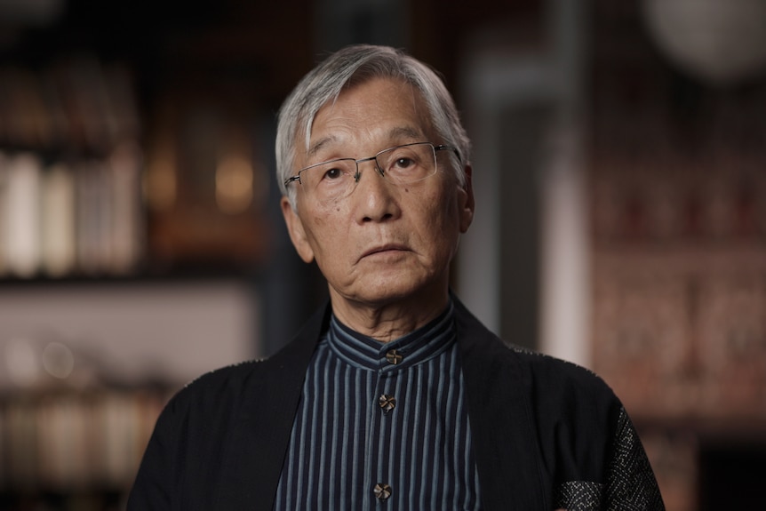 Asian man with grey hair with bookshelves behind him