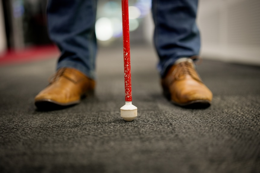 A man's feet are shown in leather shoes with a cane used by blind people resting on the floor