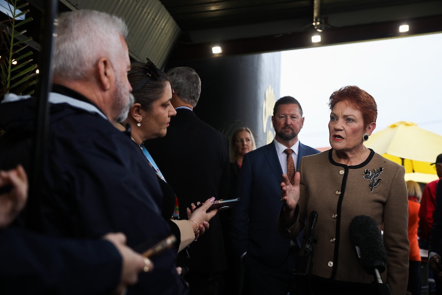 Pauline Hanson speaks to reporters among a crowd, wearing a brown and black jacket.
