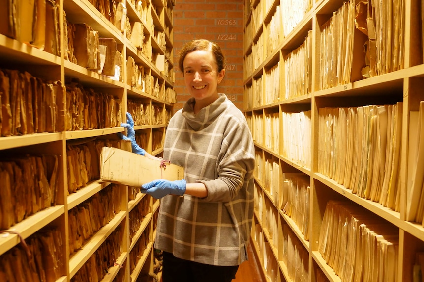 A woman stands in a tight corridor surrounded by paper files.