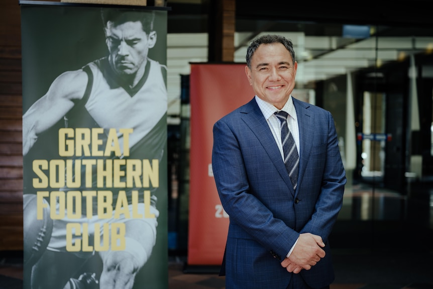 Sam smiles at the camera as he stands beside a banner featuring a footballer and the words Great Southern Football Club.