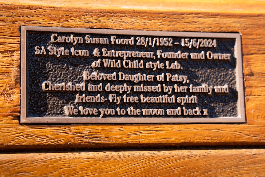 A close-up of a black memorial plaque with beige writing on a wooden bench