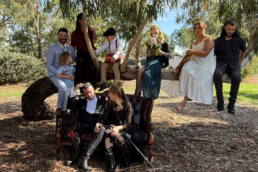 A bride and group sit on a chair in front of a tree with a group of people in formal clothes sitting on it.