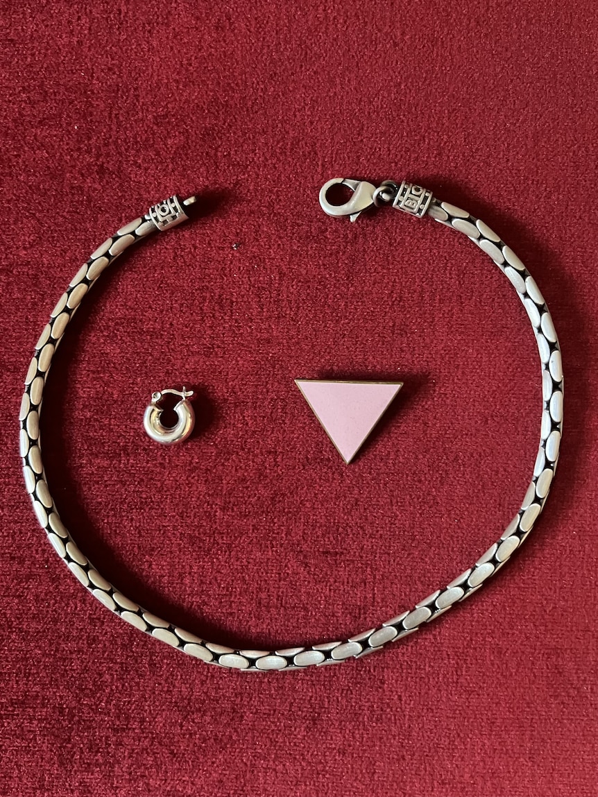 A silver chain necklace, a chunky silver sleeper earring and a pink triangle badge on a red background