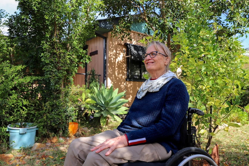 A woman sits in a wheelchair in a garden. She is smiling and wearing a blue jumper and a white scarf around her neck.