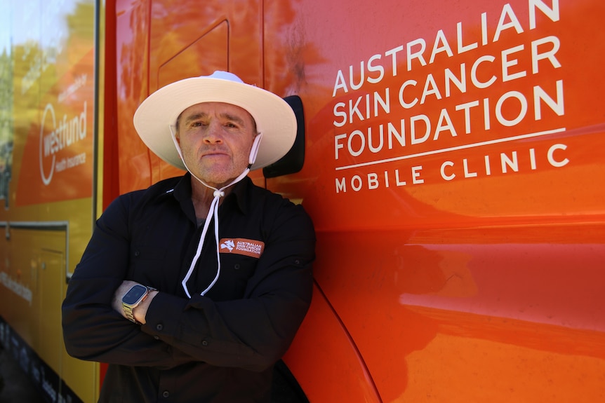 Jay Allan wearing a long sleeve black shirt with a wide brim hat leaning against orange and yellow van with arms crossed.