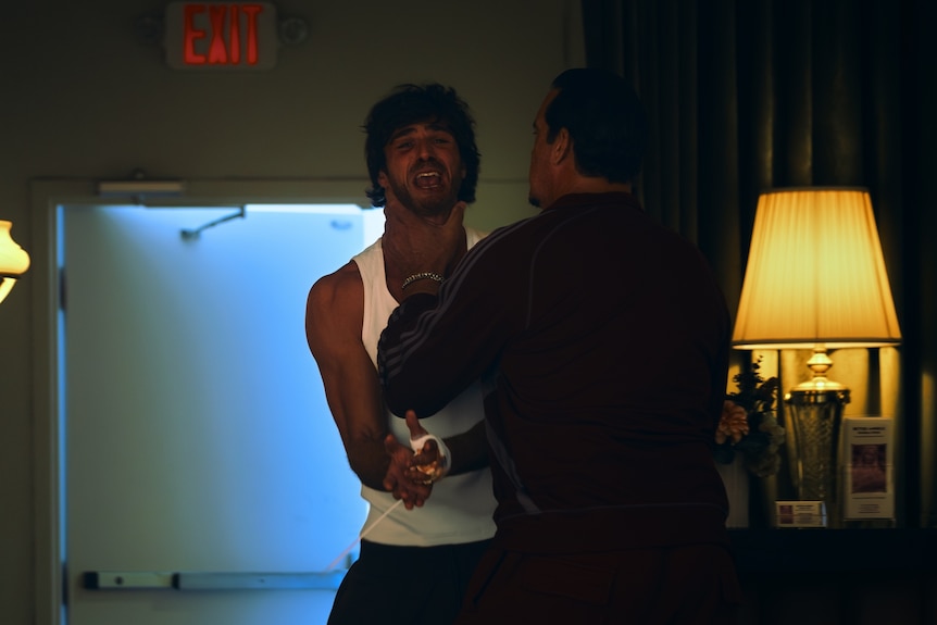Jacob Elordi with a pained expression on his face, wearing a singlet, Matthew Willig holding his neck