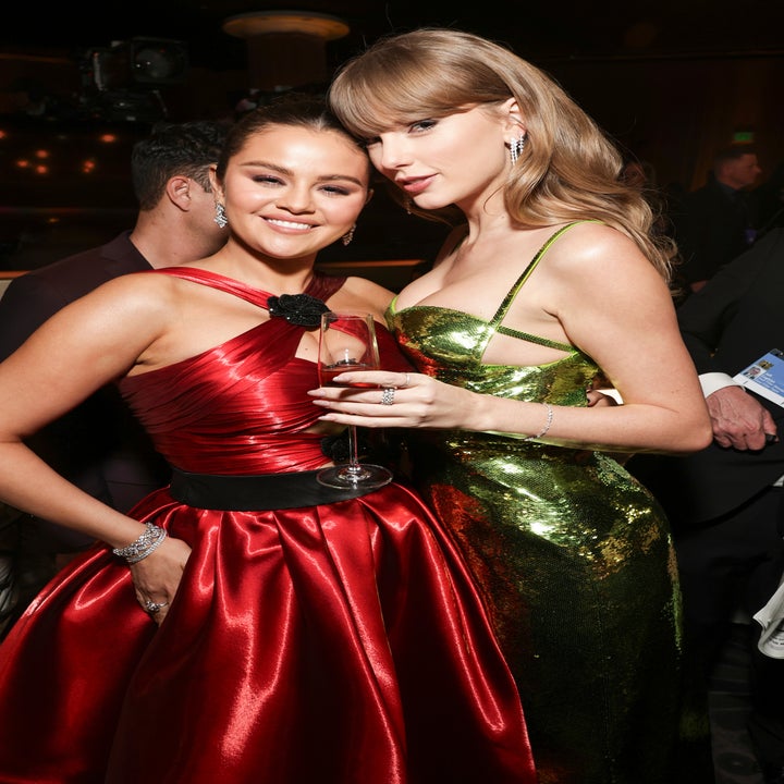 Two women in elegant, shimmering red and green dresses pose together at an event, holding a glass and smiling