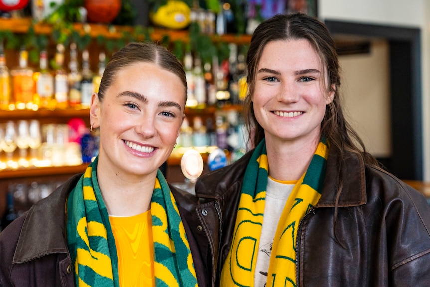 Two young women smile in a bar while wearing green and gold sports colours.