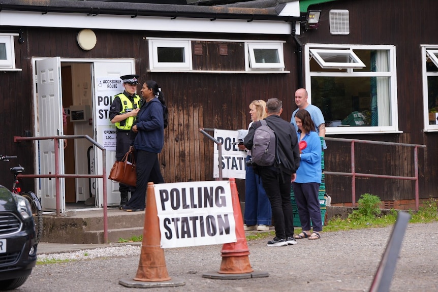 A police offiver watches on as several people prepare to enter a building that says "polling station" on it.
