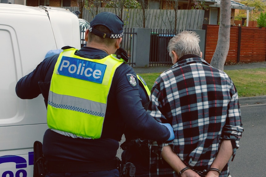 A handcuffed man is walked to the back of a police vehicle. 