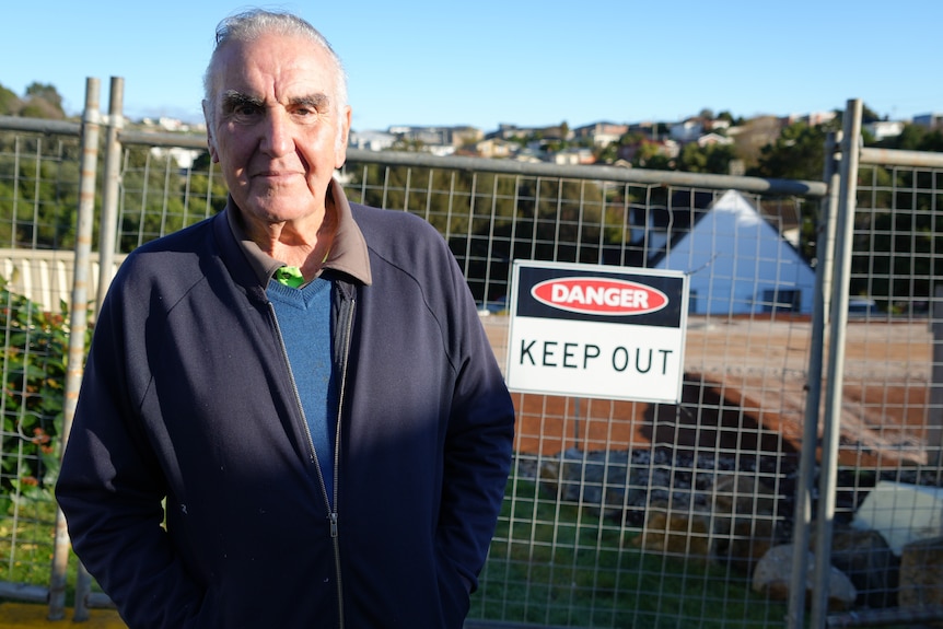 A man with grey hair and casual navy clothes stands in front of a construction site.