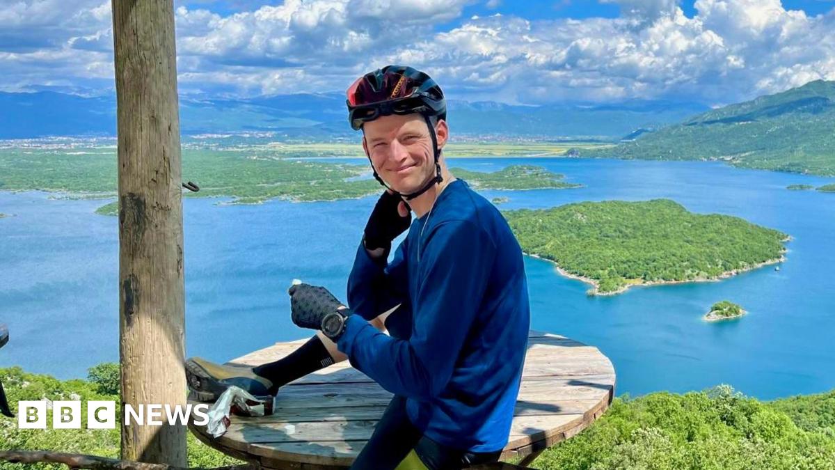 Tom Williamson in cycling clothes and a bike helmet, sitting on a wooden picnic table and smiling. A beautiful landscape with a lake, green islands and mountains is visible behind him.