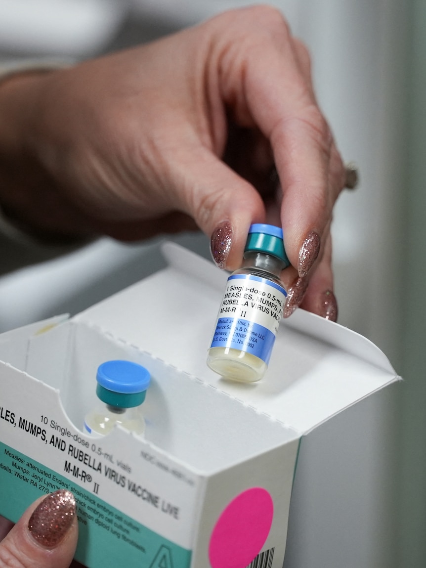 A close up of woman’s hand holding a vial of a measles, mumps and rubella vaccine out of a box.