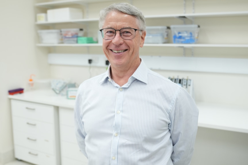 A man with short light hair and glasses stands in a lab smiling.