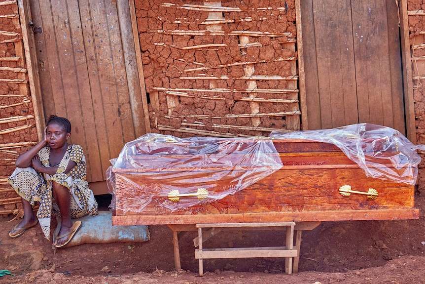 A woman sits on the ground right outside her home next to a coffin partially covered in plastic.