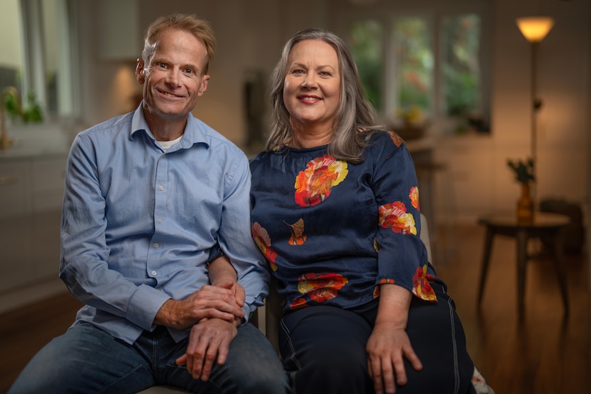 Man in a blue collared shirt seated next to a woman in dark three-quarter top, both seated and smiling