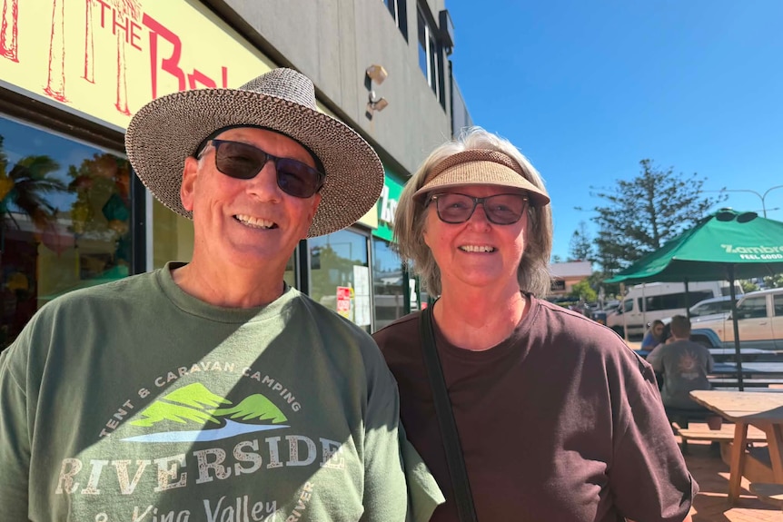 A man wearing a hat and sunglasses stand next to a woman wearing a hat and purple shirt