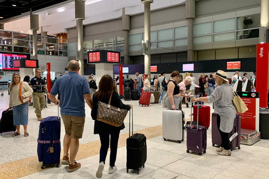 crowds of people walk through an airport with luggage, lined up at baggage drop points