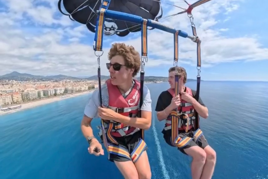 Two men hanging in seats from a paraglider in the air across the ocean.