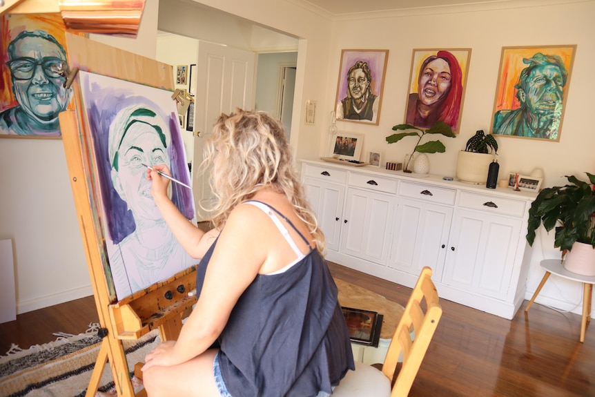 A female artist with long blonde hair sitting in front of an easel painting a portrait of a woman.