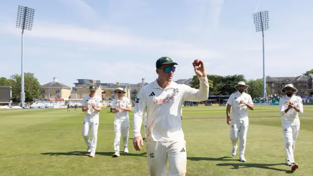 Liam Patterson-White walks off with the ball raised in his left hand after taking six wickets against Essex