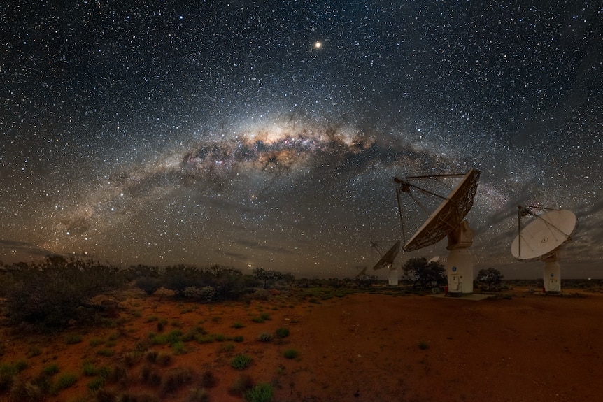 Three radio satellite dishes point at the night sky, where the milky way glows brightly, above red dirt with low scrub.