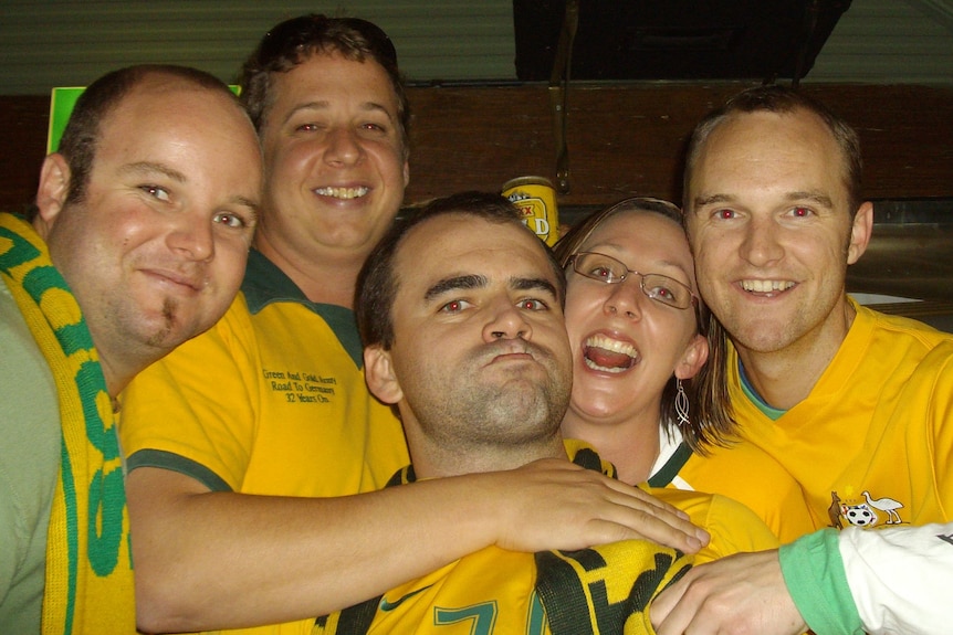 Four men and a woman, all smiling and wearing Socceroos jerseys as they cuddle up for a selfie.