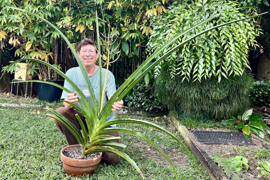 Jerry Coleby-Williams squats smiling in a backyard behind a giant potted succulent
