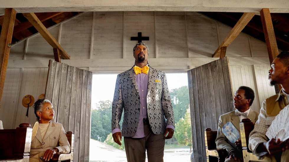 A man in a patterned suit with a yellow bow tie stands in a church, surrounded by seated people.